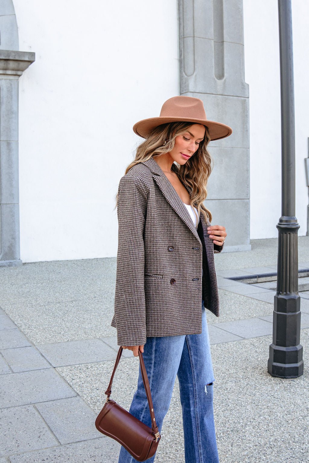 A woman wearing a brown hat and YURA YJ9036BJ BROWN DOUBLE BREASTED HOUNDSTOOTH BLAZER carries a brown shoulder bag while walking outside.