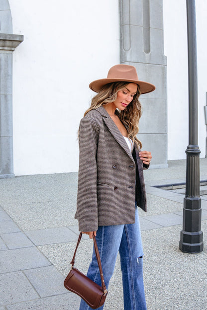 A woman wearing a brown hat and YURA YJ9036BJ BROWN DOUBLE BREASTED HOUNDSTOOTH BLAZER carries a brown shoulder bag while walking outside.
