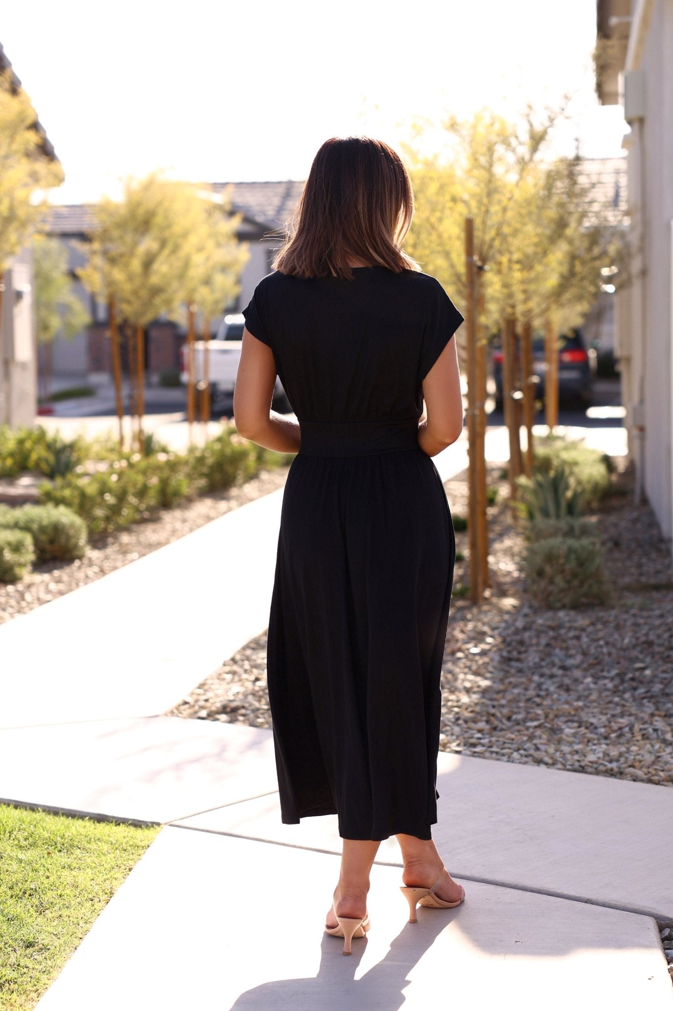 A woman in the Zoey Black Cinched Flare Midi Dress and sandals stands outside on a sunny day, facing away from the camera.
