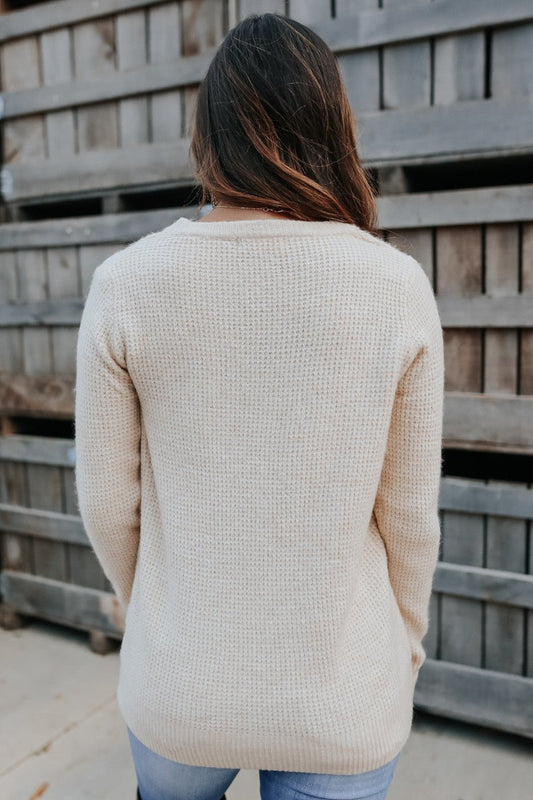 A person with long brown hair in the Autumn Glow Brushed Oatmeal Waffle Sweater and jeans stands facing a wooden fence, back to camera.