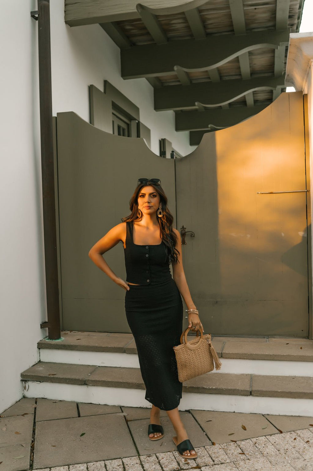 Woman in the Black Sheer Lace Midi Skirt - FINAL SALE, with sandals and woven bag, stands on steps before a gray gate at sunset.