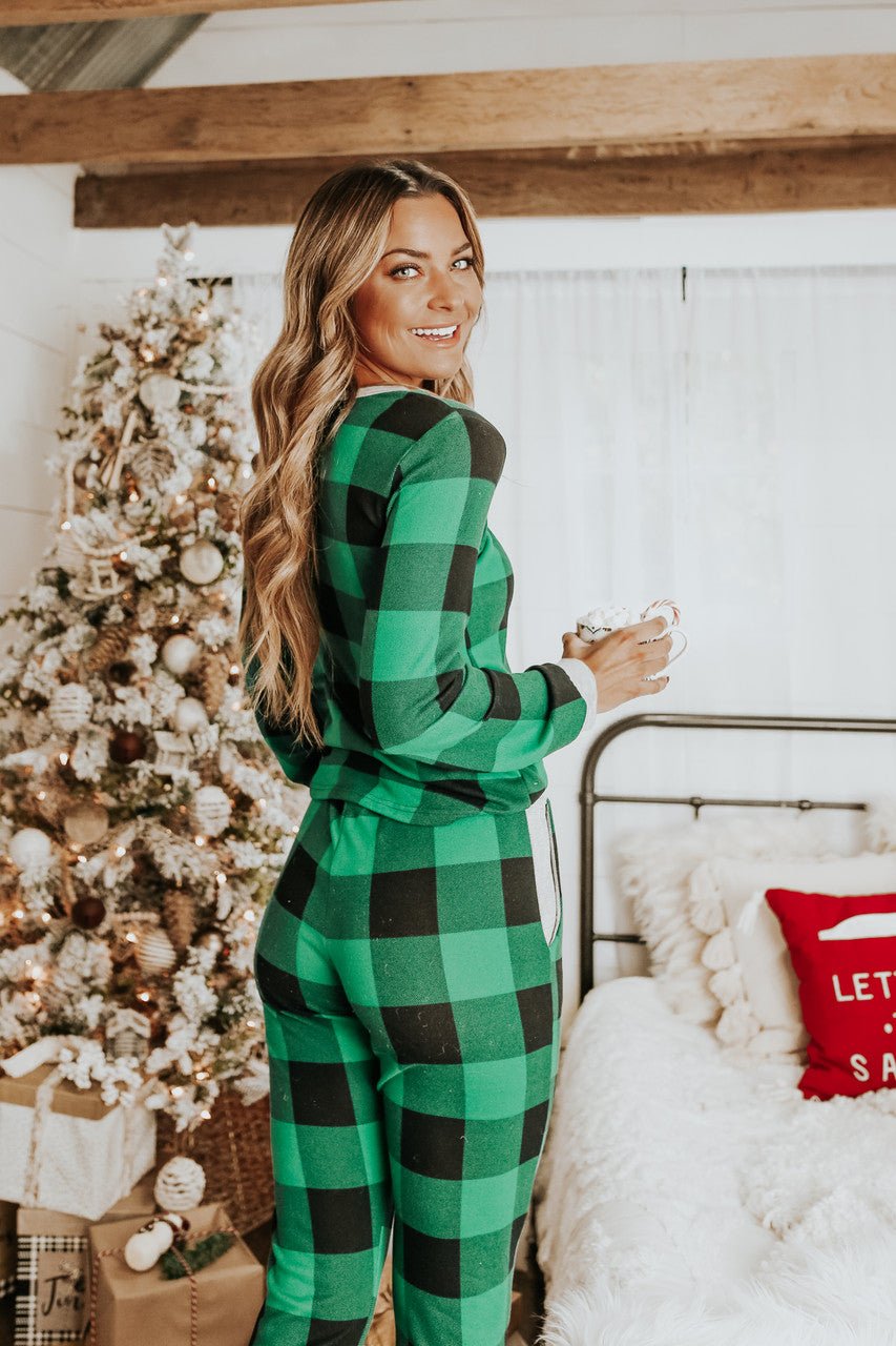 Woman in the Christmas Morning Green Plaid Pajama Set holds a mug by a festive tree and bed.