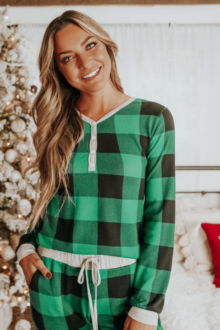 A woman in the Christmas Morning Green Plaid Pajama Set stands by a tree, with white curtains and a red pillow behind her.