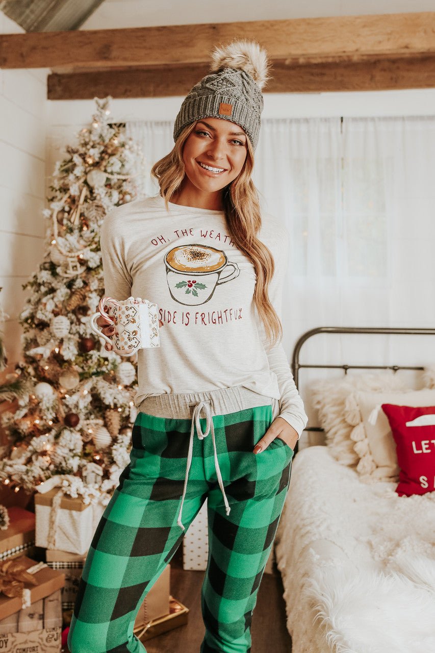 A woman in the Christmas Morning Green Plaid Pajama Set and a gray beanie holds a mug by a festive tree and bed.
