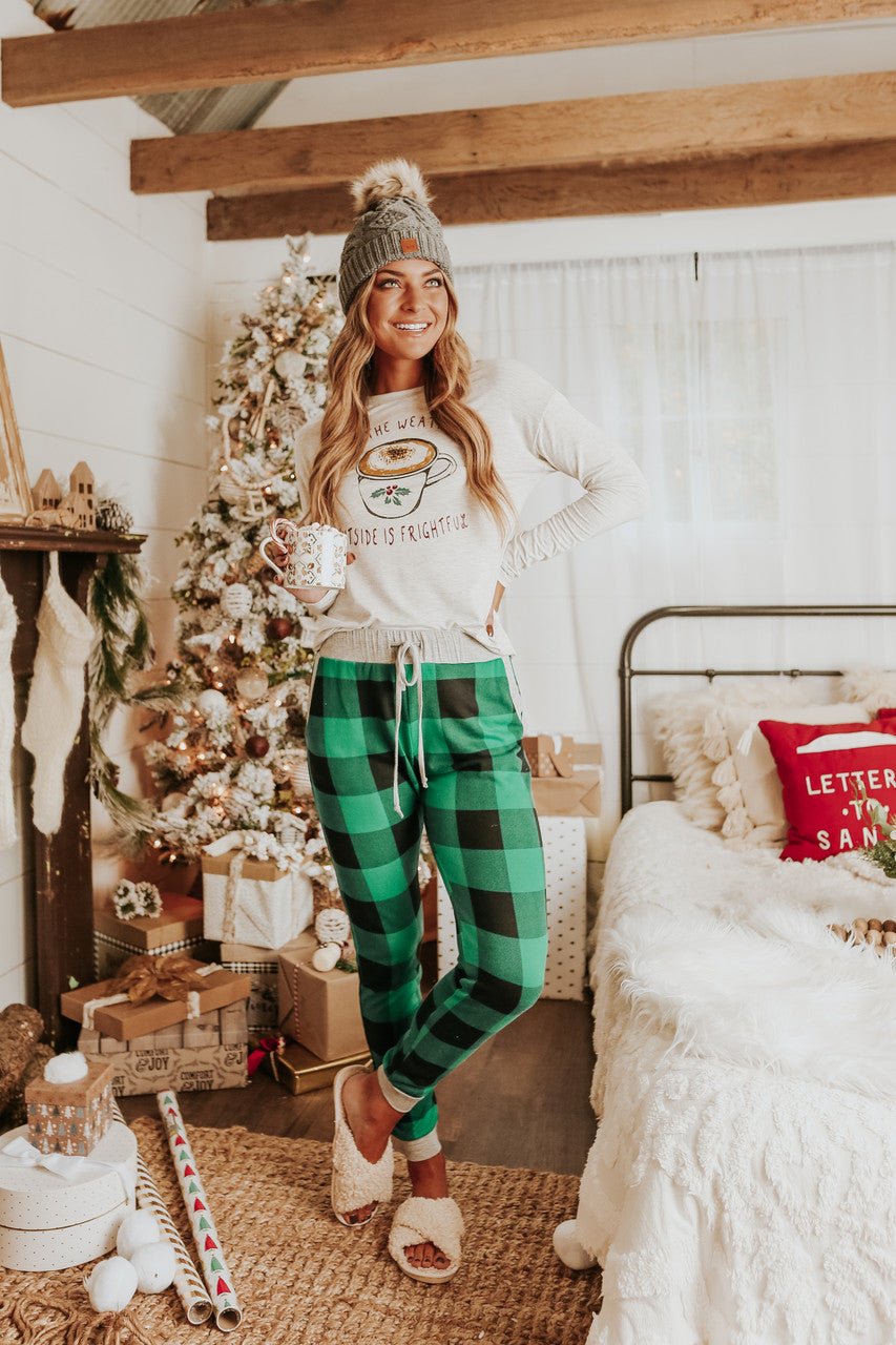 Woman wears Christmas Morning Green Plaid Pajama Set, holding a mug by a decorated tree and bed in a festive room.