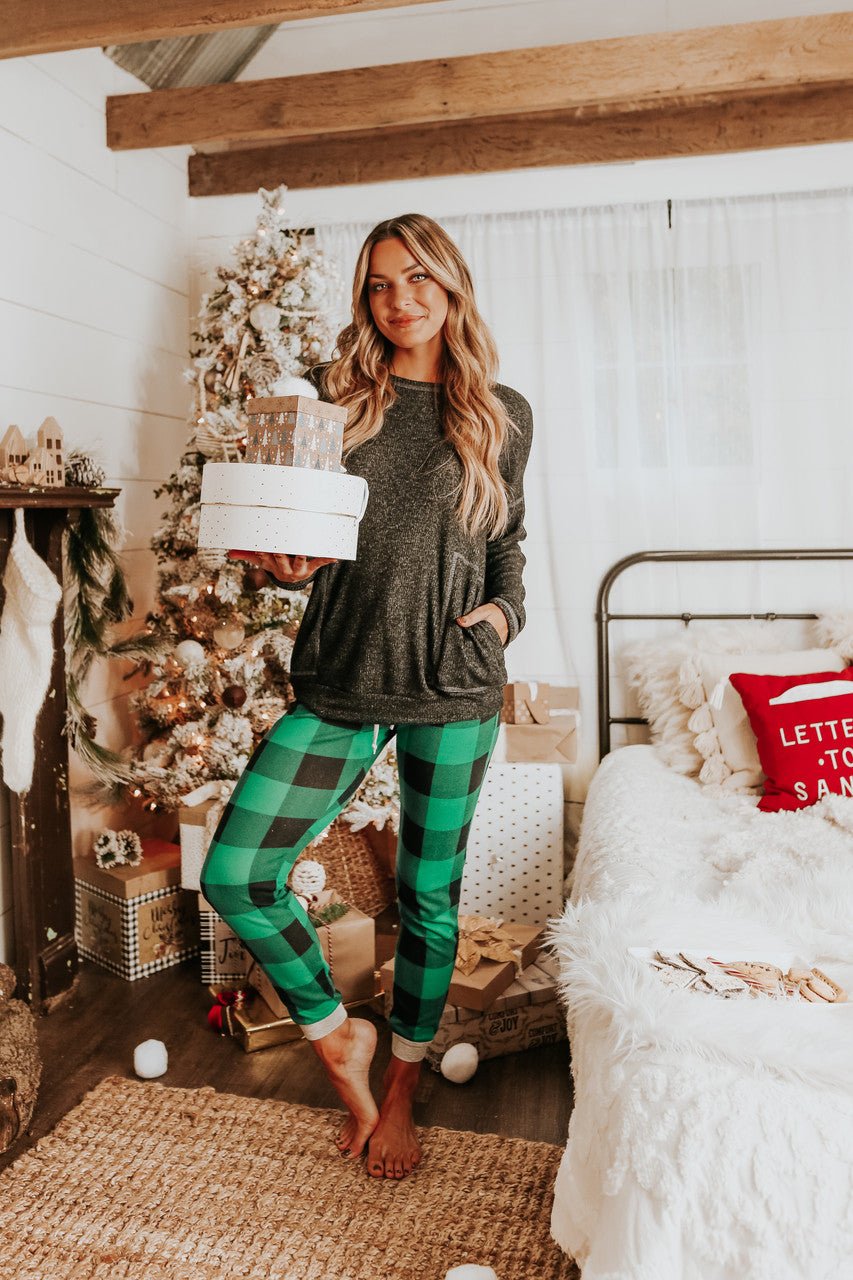 A woman in the Christmas Morning Green Plaid Pajama Set stands by a festive tree, holding a gift in her cozy holiday room.