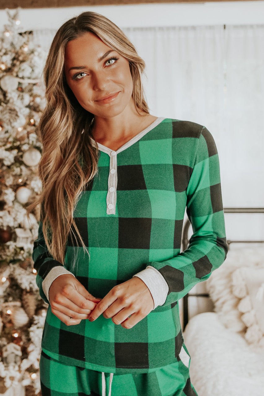 A woman wears the Christmas Morning Green Plaid Pajama Set indoors by a festive tree, smiling softly.