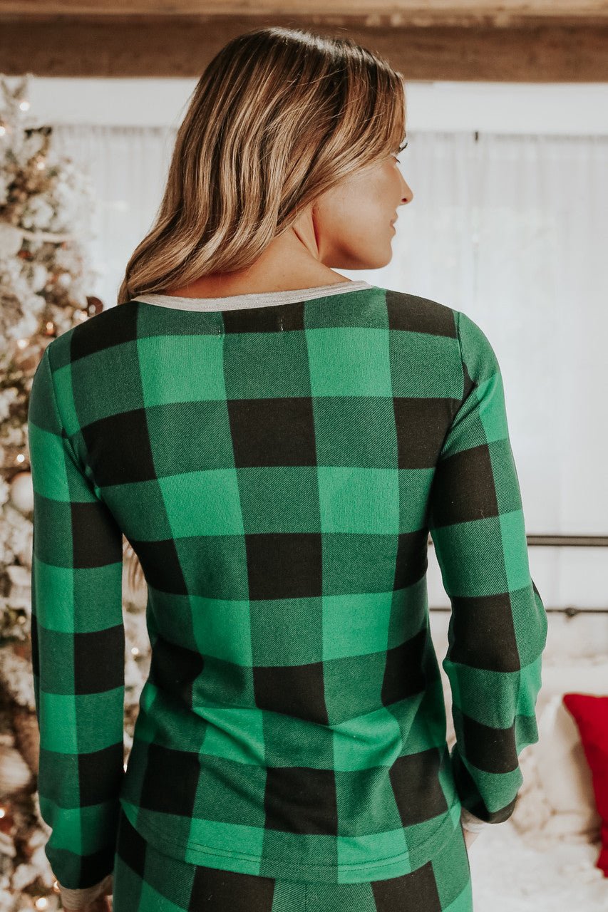 A woman with wavy hair in the Christmas Morning Green Plaid Pajama Set stands indoors by a decorated Christmas tree.
