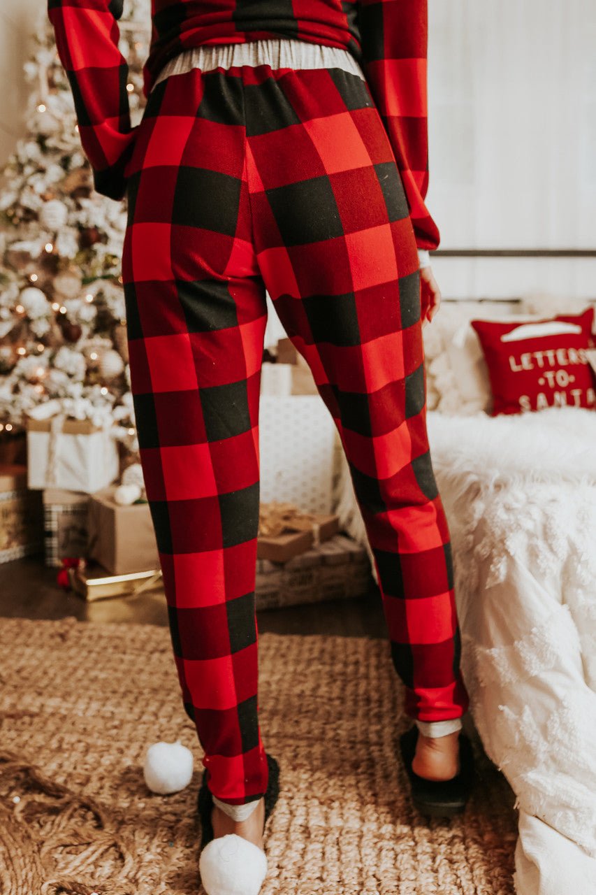 Wearing the Christmas Morning Red Plaid Pajama Set, a person stands by a festively decorated bed and tree, ready for Christmas morning.