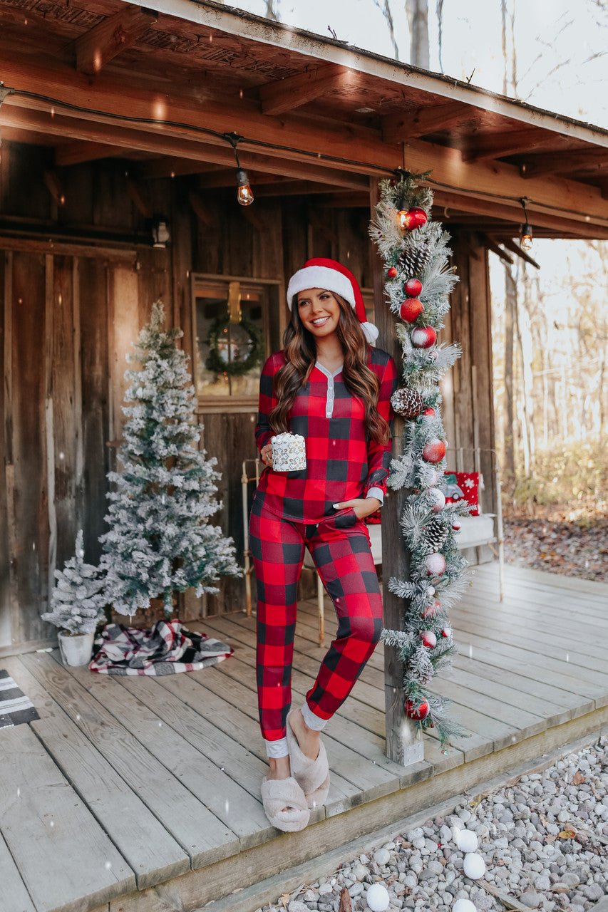 Woman in the Christmas Morning Red Plaid Pajama Set stands on a festive porch with a mug, smiling by decorated Christmas trees.