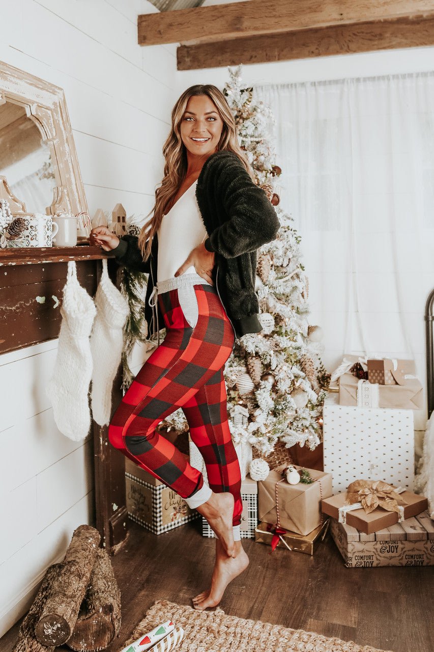 Woman in the Christmas Morning Red Plaid Pajama Set stands by a festive tree and fireplace, surrounded by wrapped gifts.