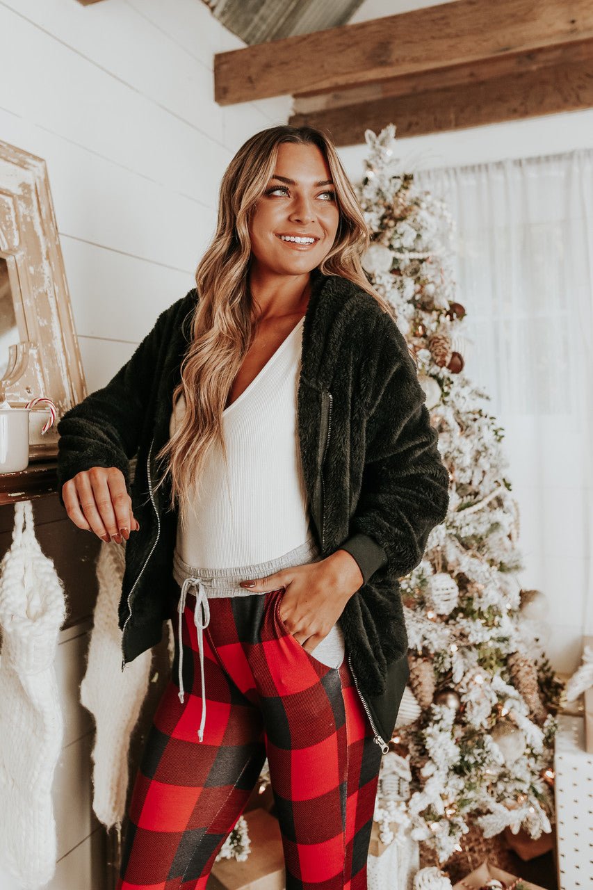 Woman smiles by a Christmas tree, wearing the Christmas Morning Red Plaid Pajama Set in a festive, rustic room.