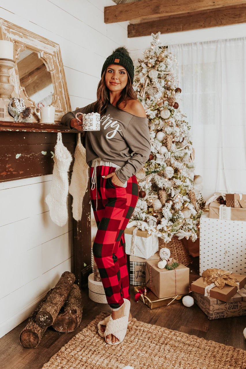 Woman in Christmas Morning Red Plaid Pajama Set holds a mug by a decorated tree, fireplace, and wrapped gifts.