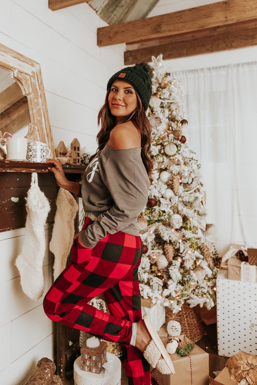 A woman in the Christmas Morning Red Plaid Pajama Set smiles by a festive tree and fireplace in a cozy holiday scene.