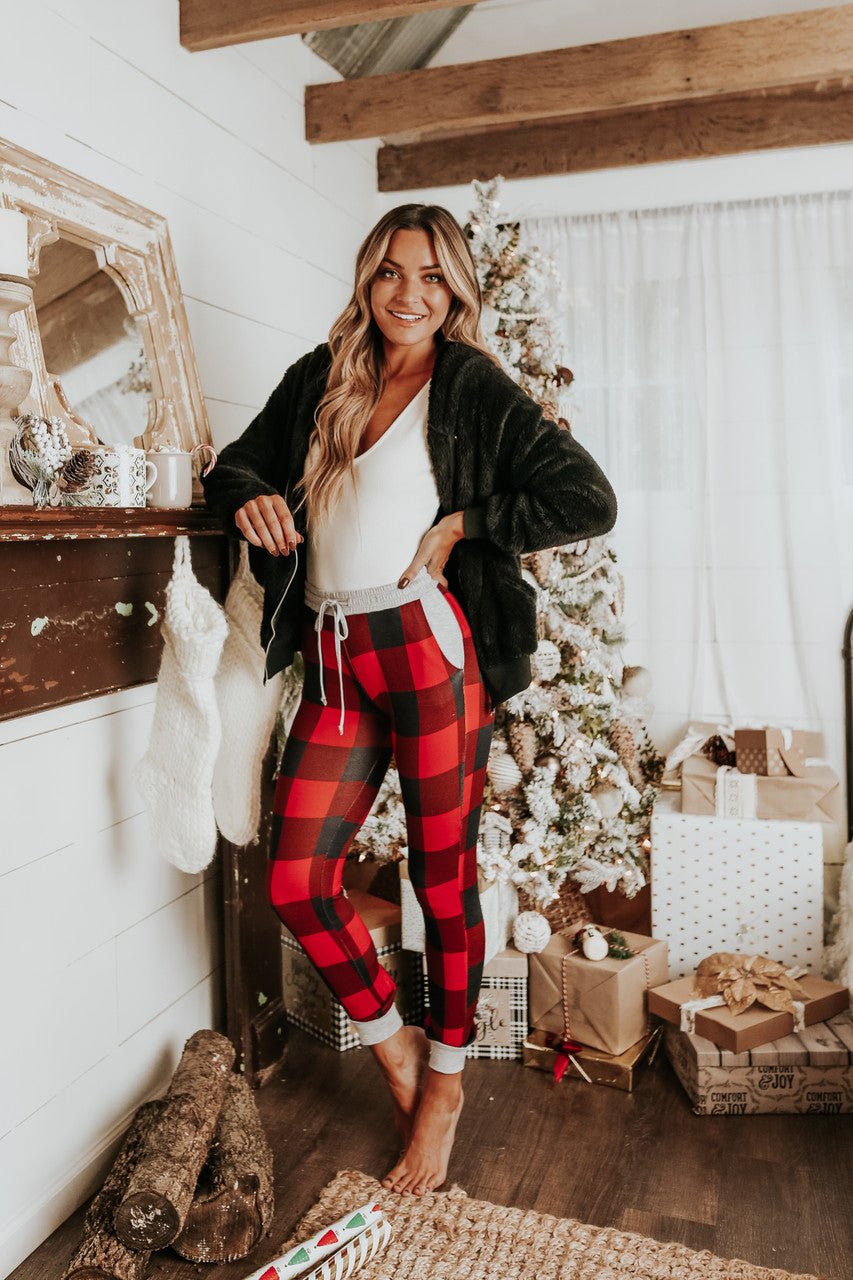A woman in a Christmas Morning Red Plaid Pajama Set stands barefoot by a decorated tree, fireplace, gifts, and stockings.