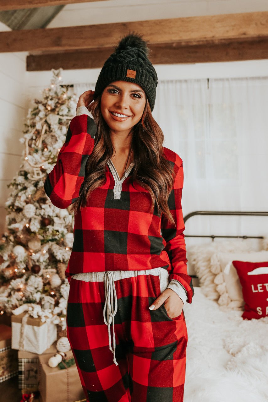 A woman in a Christmas Morning Red Plaid Pajama Set and black beanie smiles by a festive tree in a cozy holiday bedroom.
