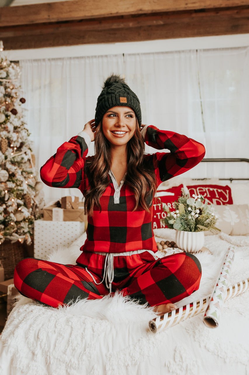 Woman in a red plaid Christmas Morning Red Plaid Pajama Set sits cross-legged on a bed by the tree, smiling.