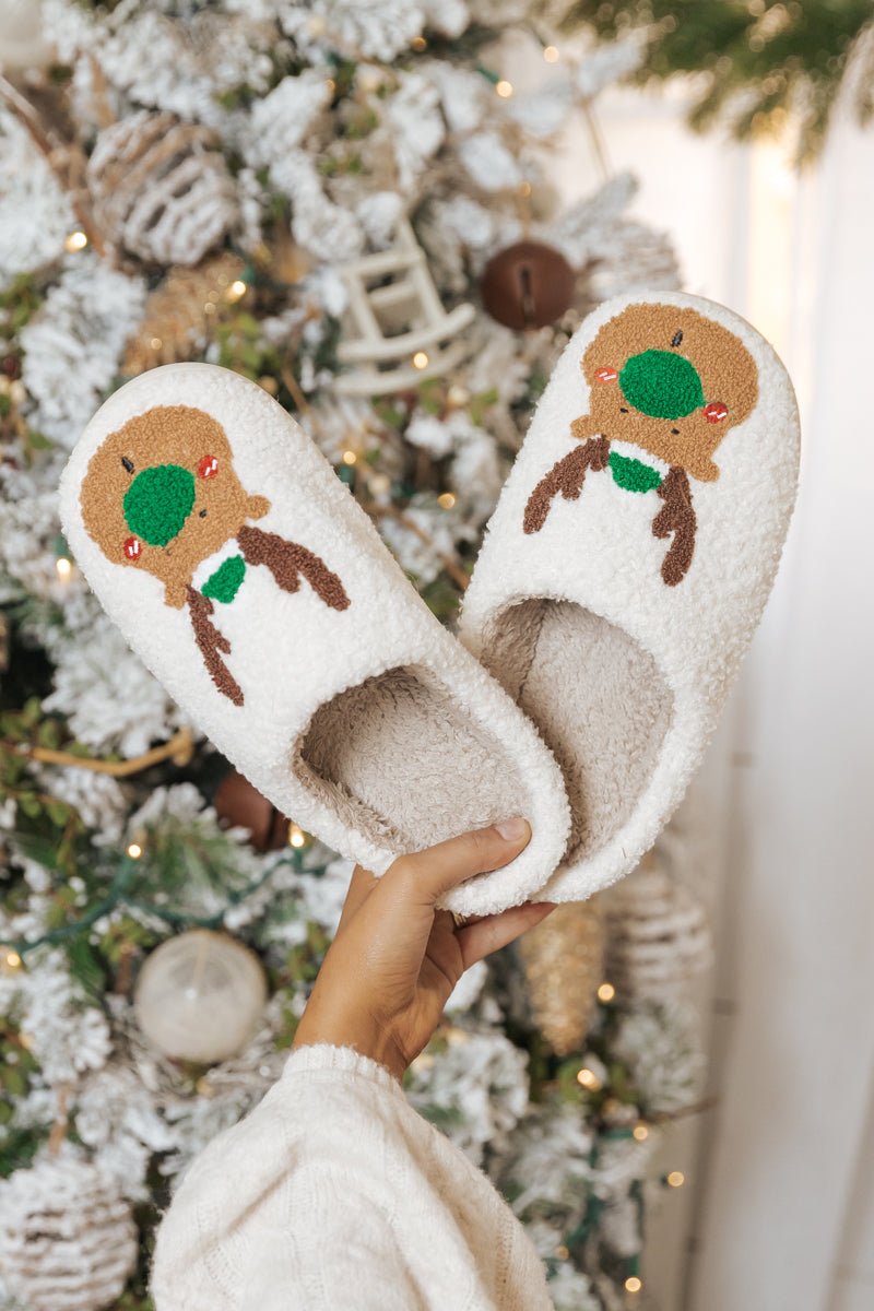 A hand holds Cream Faux Plush Reindeer Slippers—FINAL SALE—in front of a festive, snow-decorated Christmas tree; great holiday gift!.