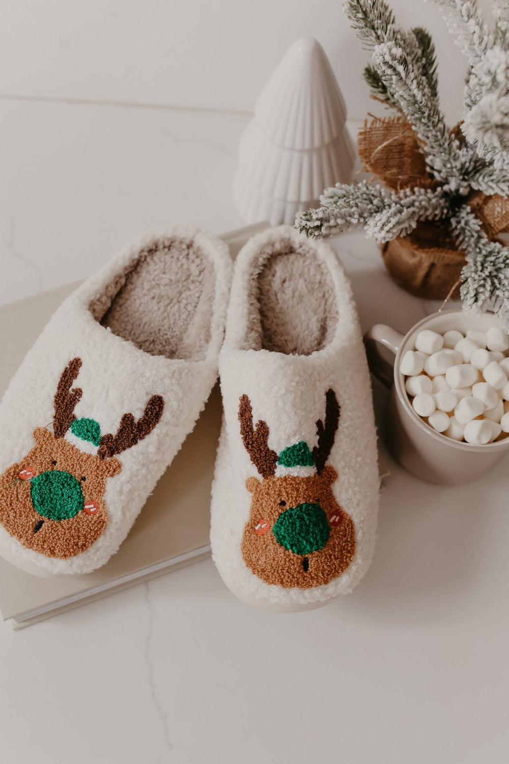 Cream Faux Plush Reindeer Slippers sit by a marshmallow mug and frosted pine—an ideal Christmas gift on a white surface.