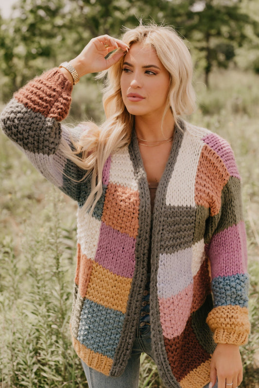 Woman in Falling Leaves Olive Color Block Cardigan stands outdoors, touching her hair, greenery behind her.