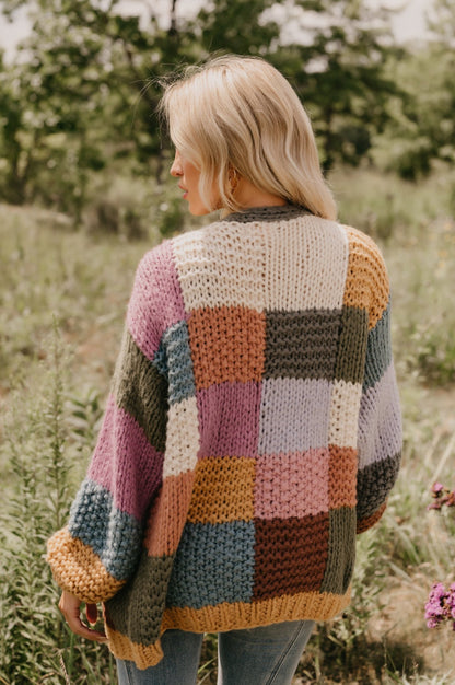 A person with blonde hair wears the Falling Leaves Olive Color Block Cardigan while standing in a grassy field.