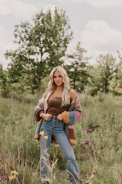 A woman stands in a grassy field wearing jeans, a brown top, and the Falling Leaves Olive Color Block Cardigan - FINAL SALE.