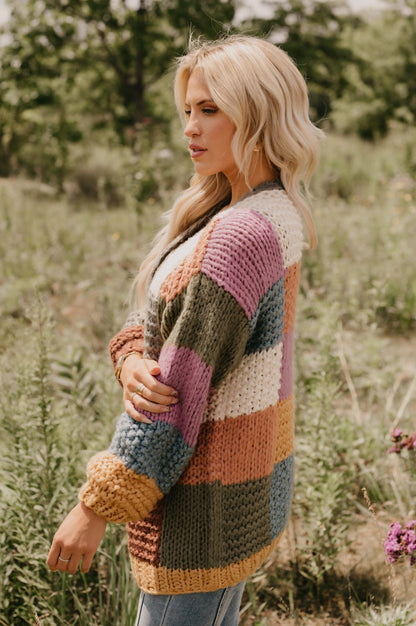 A woman with long blonde hair wears the Falling Leaves Olive Color Block Cardigan while standing outdoors in a grassy field.