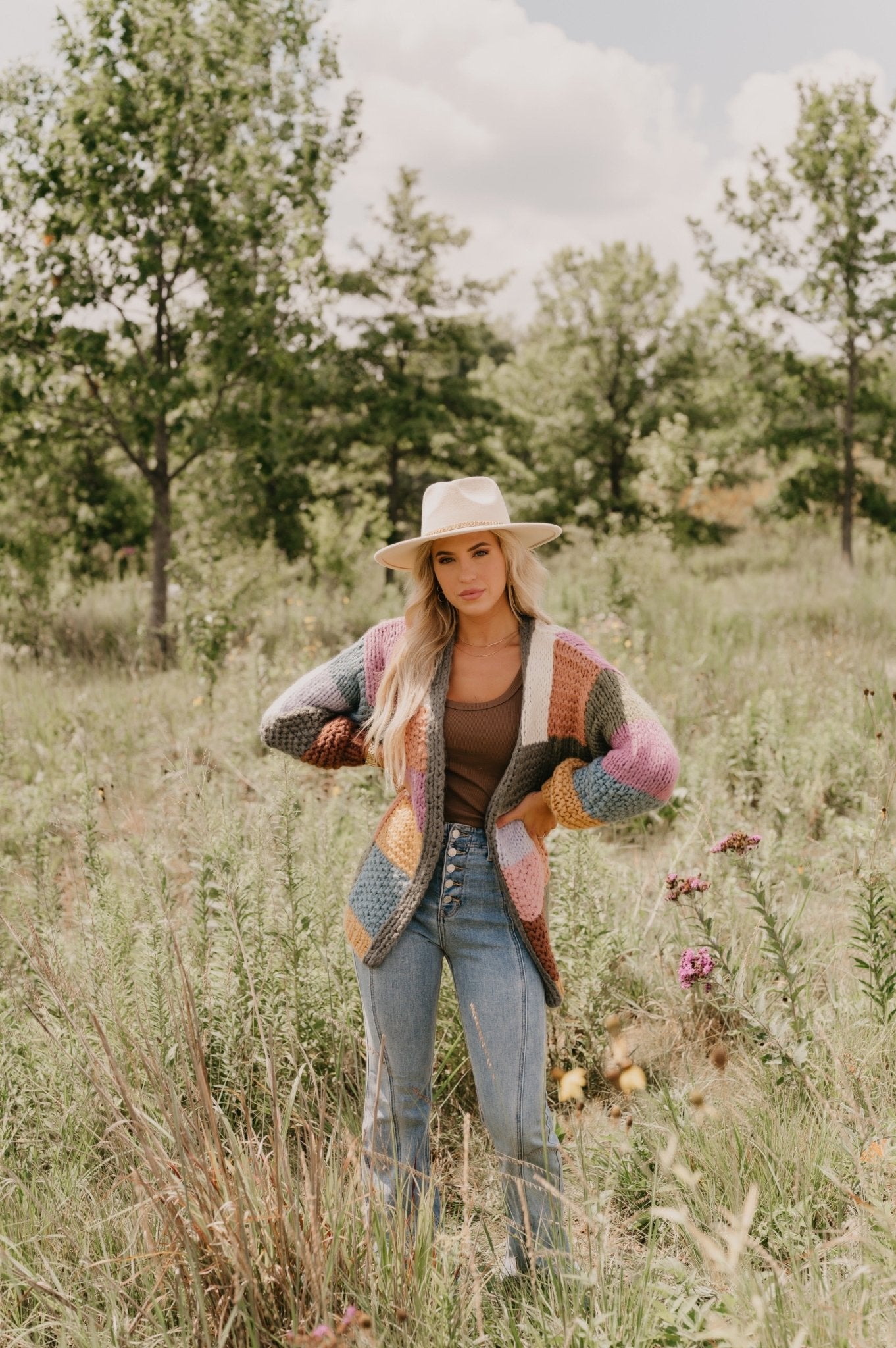 Woman in the Falling Leaves Olive Color Block Cardigan stands in a grassy field with wildflowers and green trees behind her.