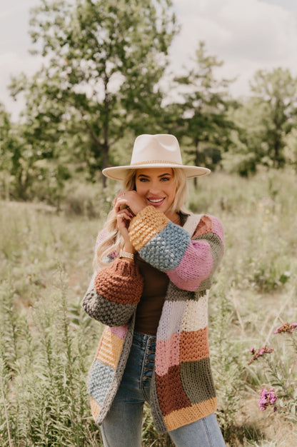 A smiling woman wears the Falling Leaves Olive Color Block Cardigan, standing in a grassy field with trees behind her.