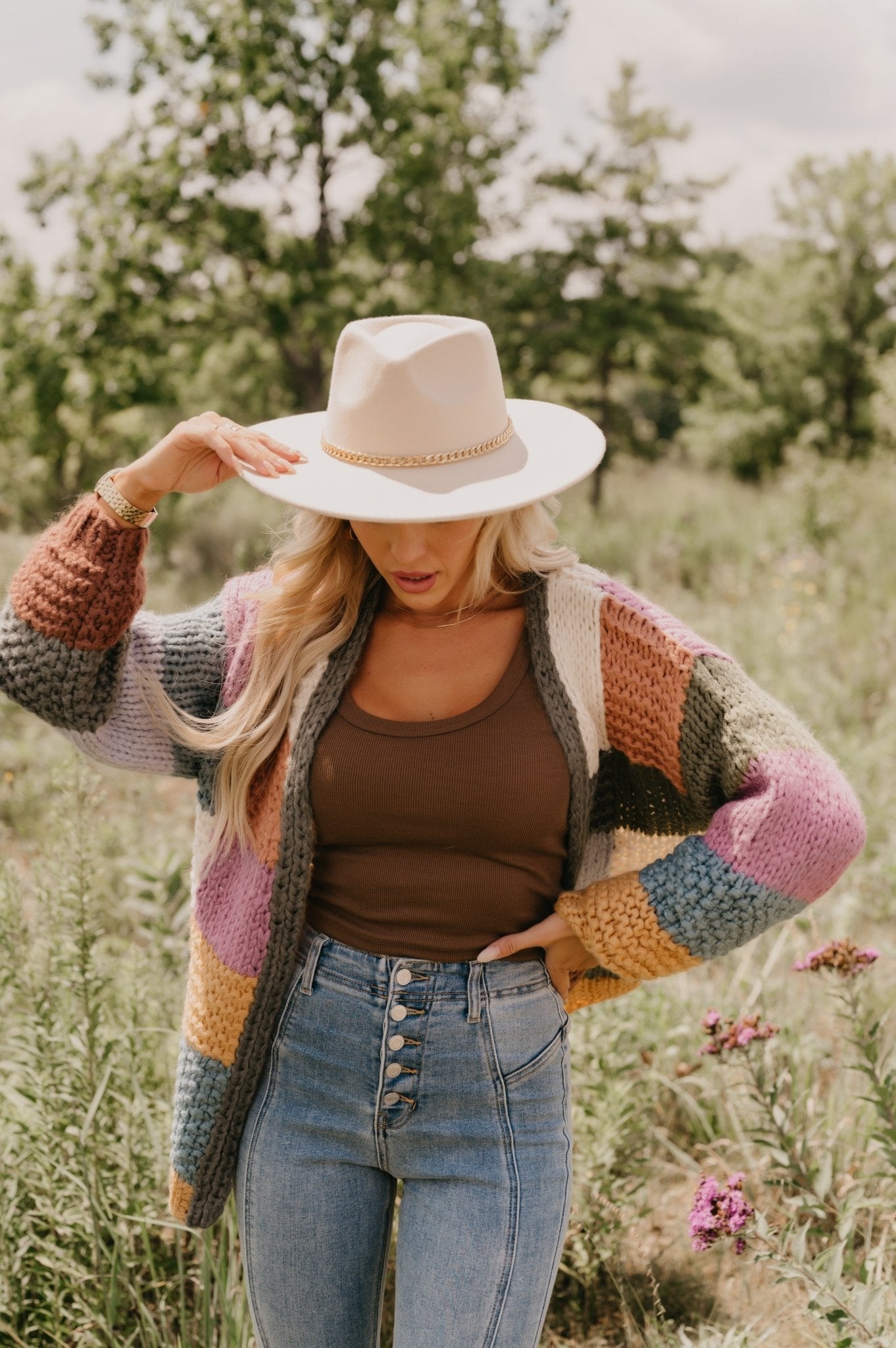 A woman in the Falling Leaves Olive Color Block Cardigan stands outdoors among greenery and wildflowers.