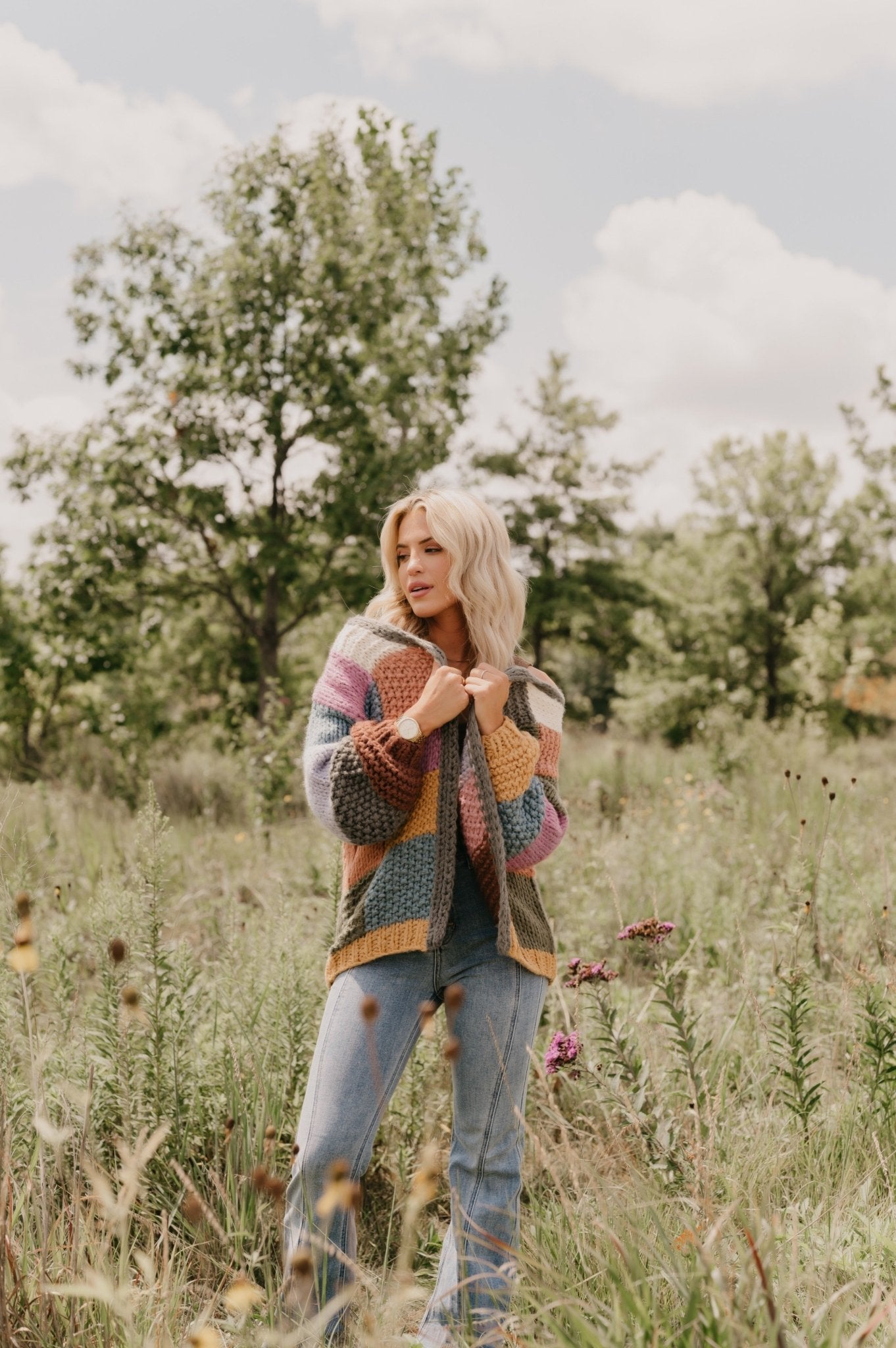 Woman with blonde hair wears the Falling Leaves Olive Color Block Cardigan and jeans in a grassy field with trees.