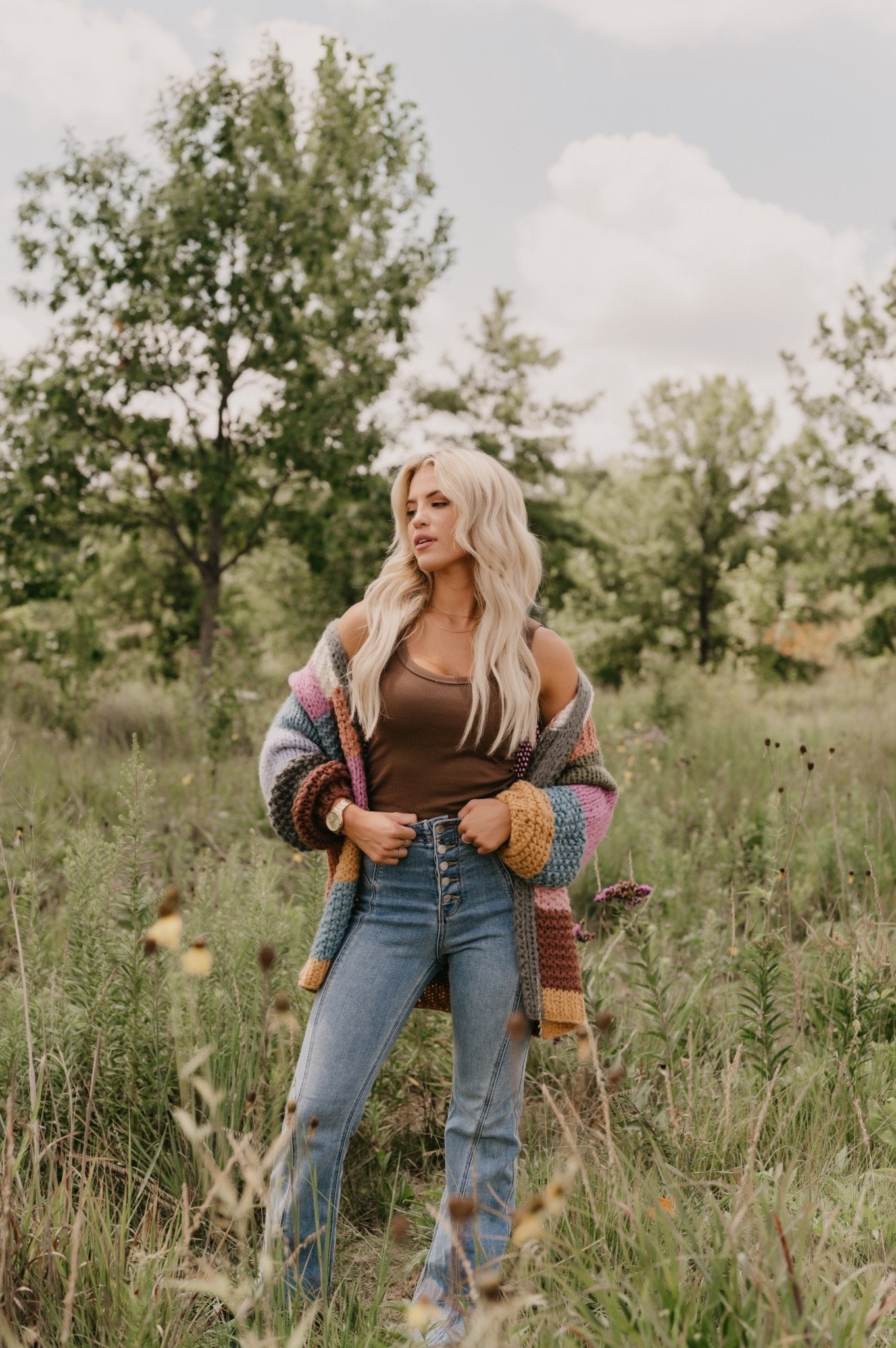 A woman with long blond hair stands in a grassy field wearing jeans, a brown top, and the Falling Leaves Olive Color Block Cardigan.