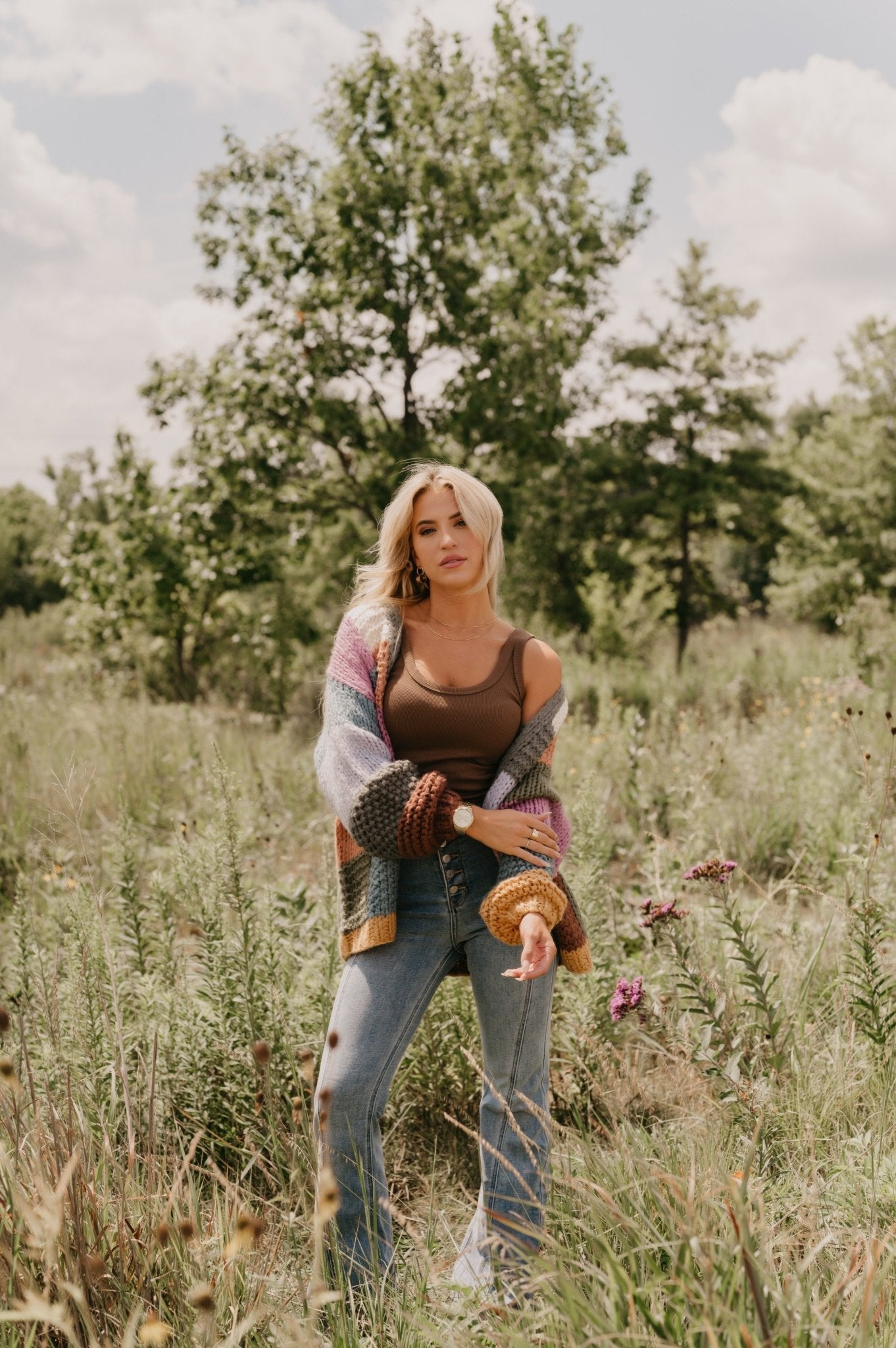 A woman in the Falling Leaves Olive Color Block Cardigan stands in a grassy field with trees and blue sky behind her.