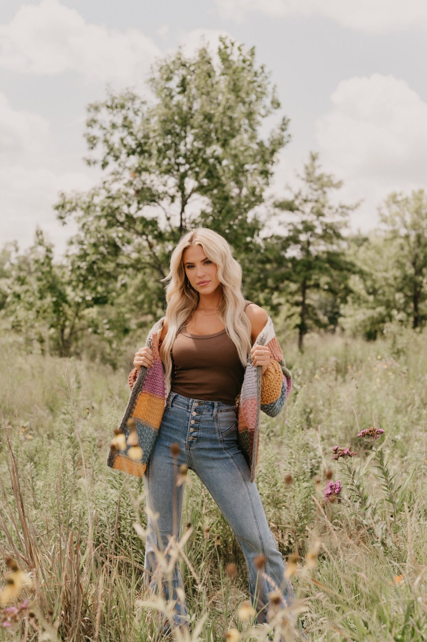 A blonde woman in the Falling Leaves Olive Color Block Cardigan and jeans stands in a grassy field on a partly cloudy day.