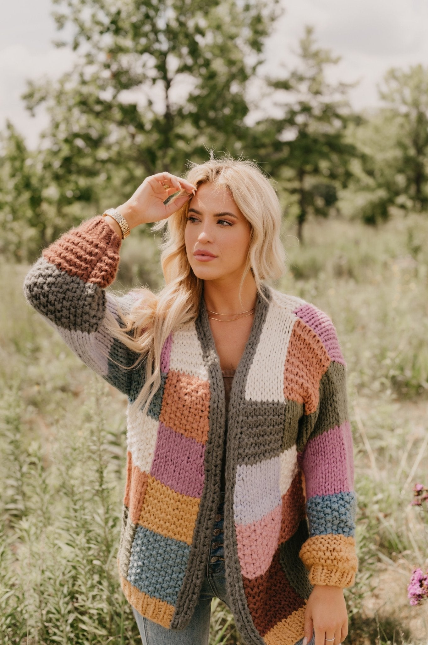 A woman wears the Falling Leaves Olive Color Block Cardigan, standing outdoors in a grassy area and touching her hair.