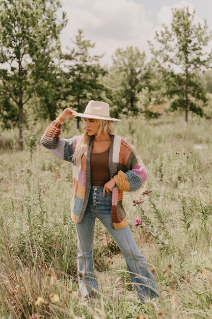 A woman wears the Falling Leaves Olive Color Block Cardigan in a grassy field with trees in the background.