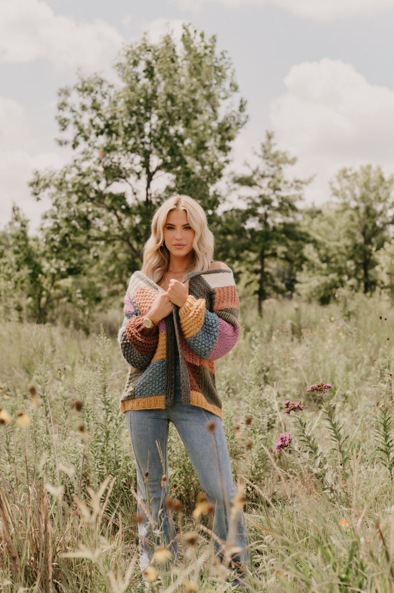 A woman wears the Falling Leaves Olive Color Block Cardigan and jeans in a grassy field with wildflowers and trees.