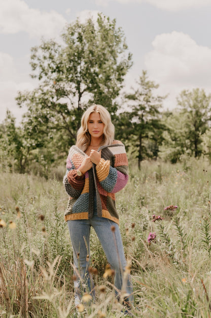 A woman wears the Falling Leaves Olive Color Block Cardigan and jeans in a grassy field with wildflowers and trees.