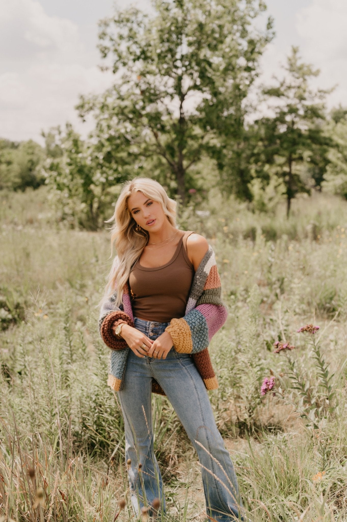 Woman with long blonde hair wears the Falling Leaves Olive Color Block Cardigan, brown tank top, and jeans in a grassy field.