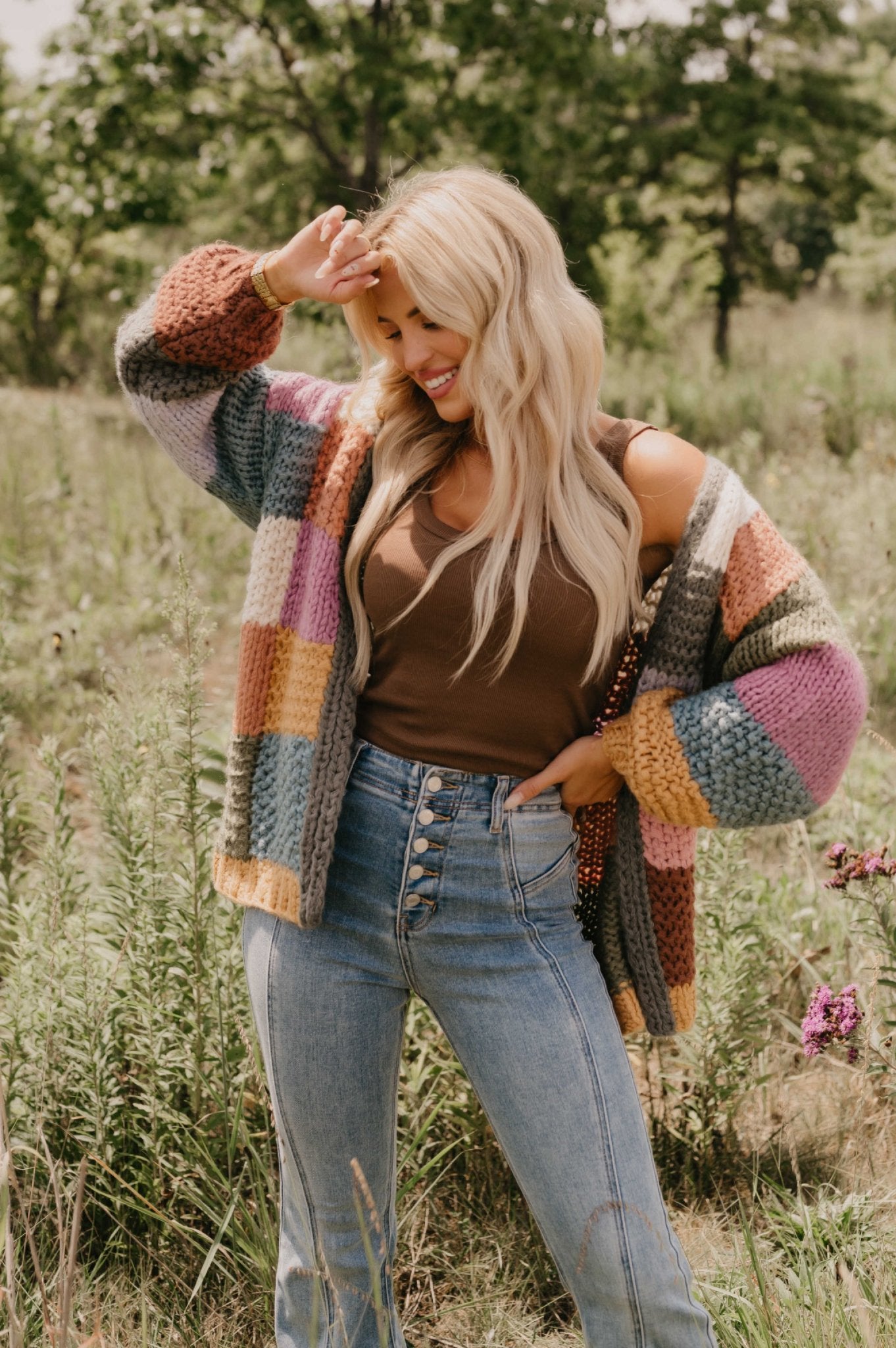 Smiling woman with long blonde hair wears the Falling Leaves Olive Color Block Cardigan and jeans outdoors in a sunny, grassy field.