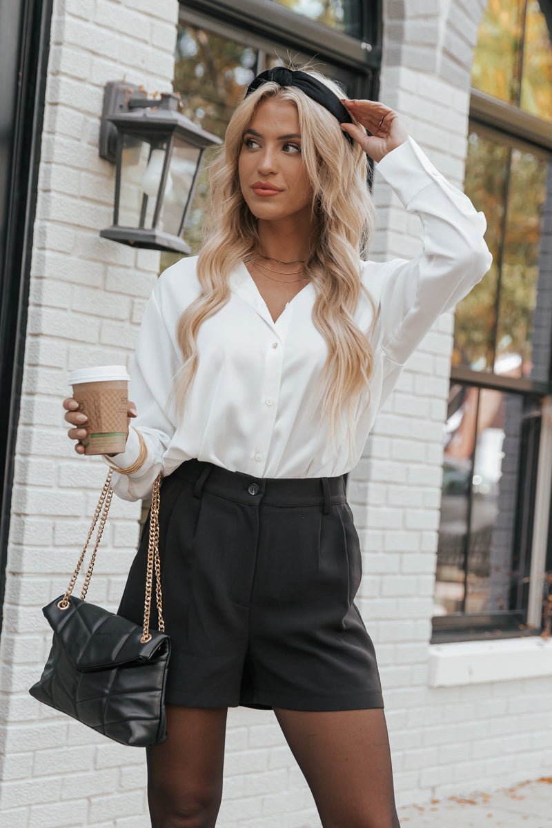 Woman in a white blouse and Muse by Magnolia Black Pleated Detail Shorts holds coffee and a black purse by a white brick wall.