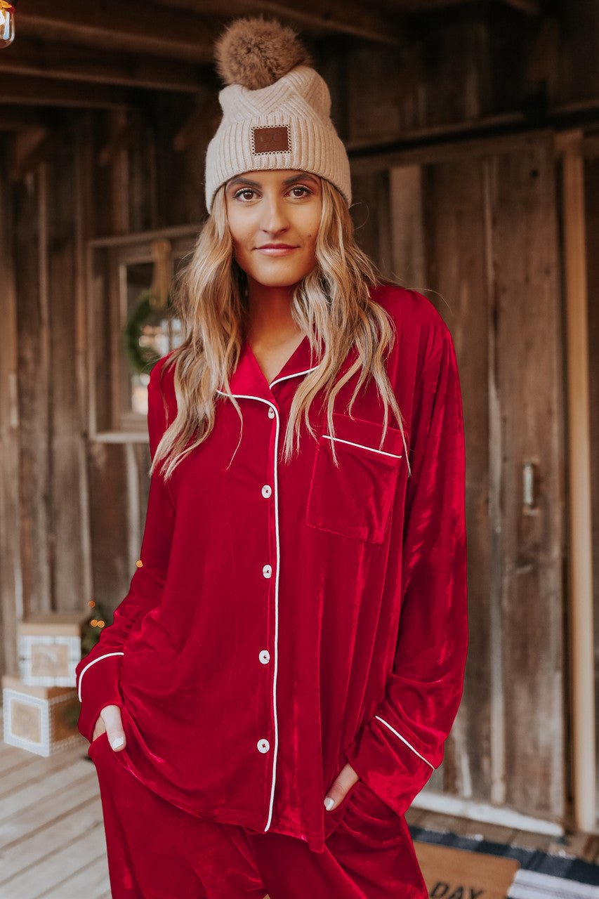 A woman in a North Pole Burgundy Velvet Pajama Set stands on a rustic porch, smiling softly at the camera.
