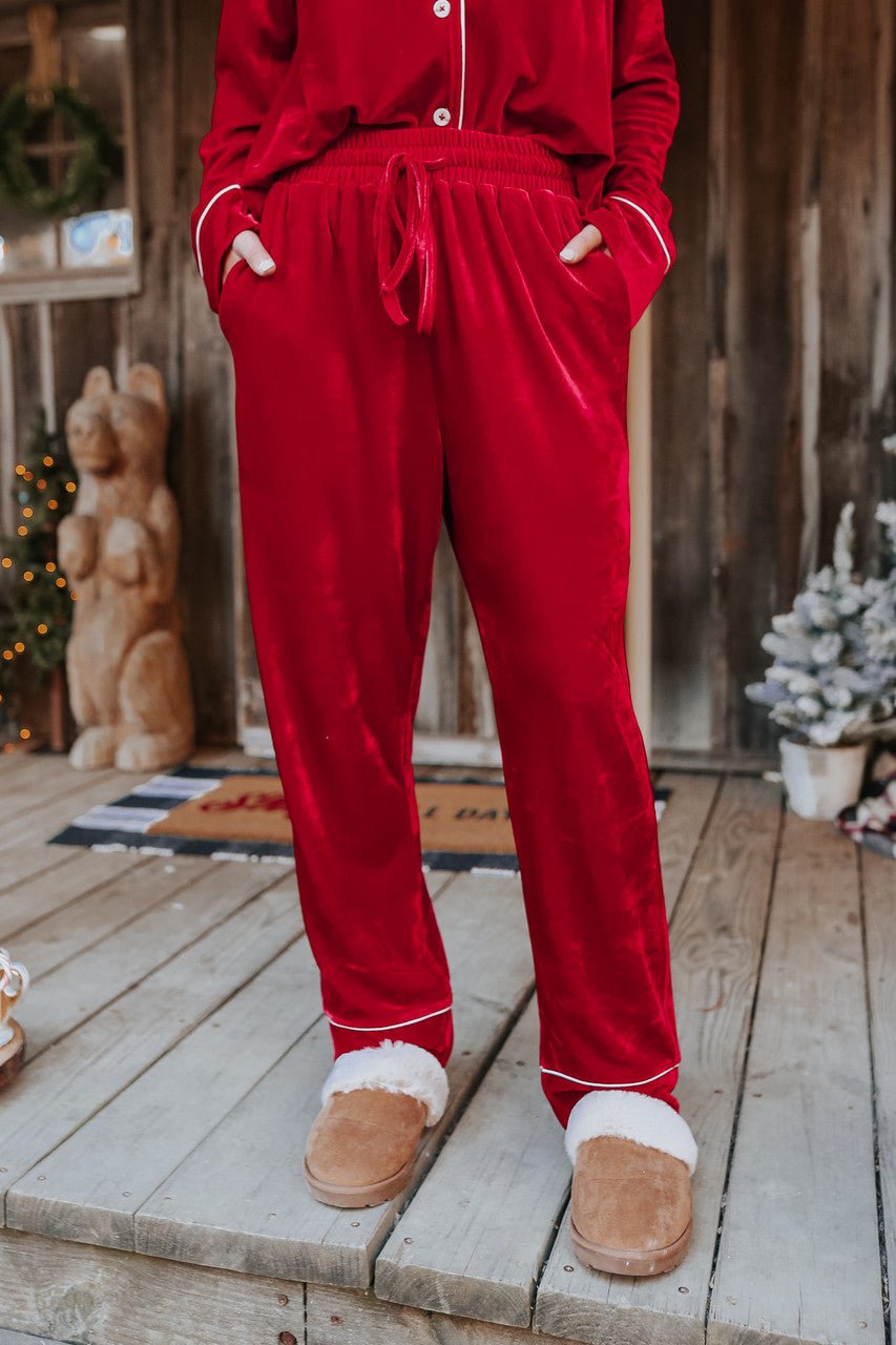 Person in the North Pole Burgundy Velvet Pajama Set stands on a porch decorated with holiday ornaments and a small Christmas tree.
