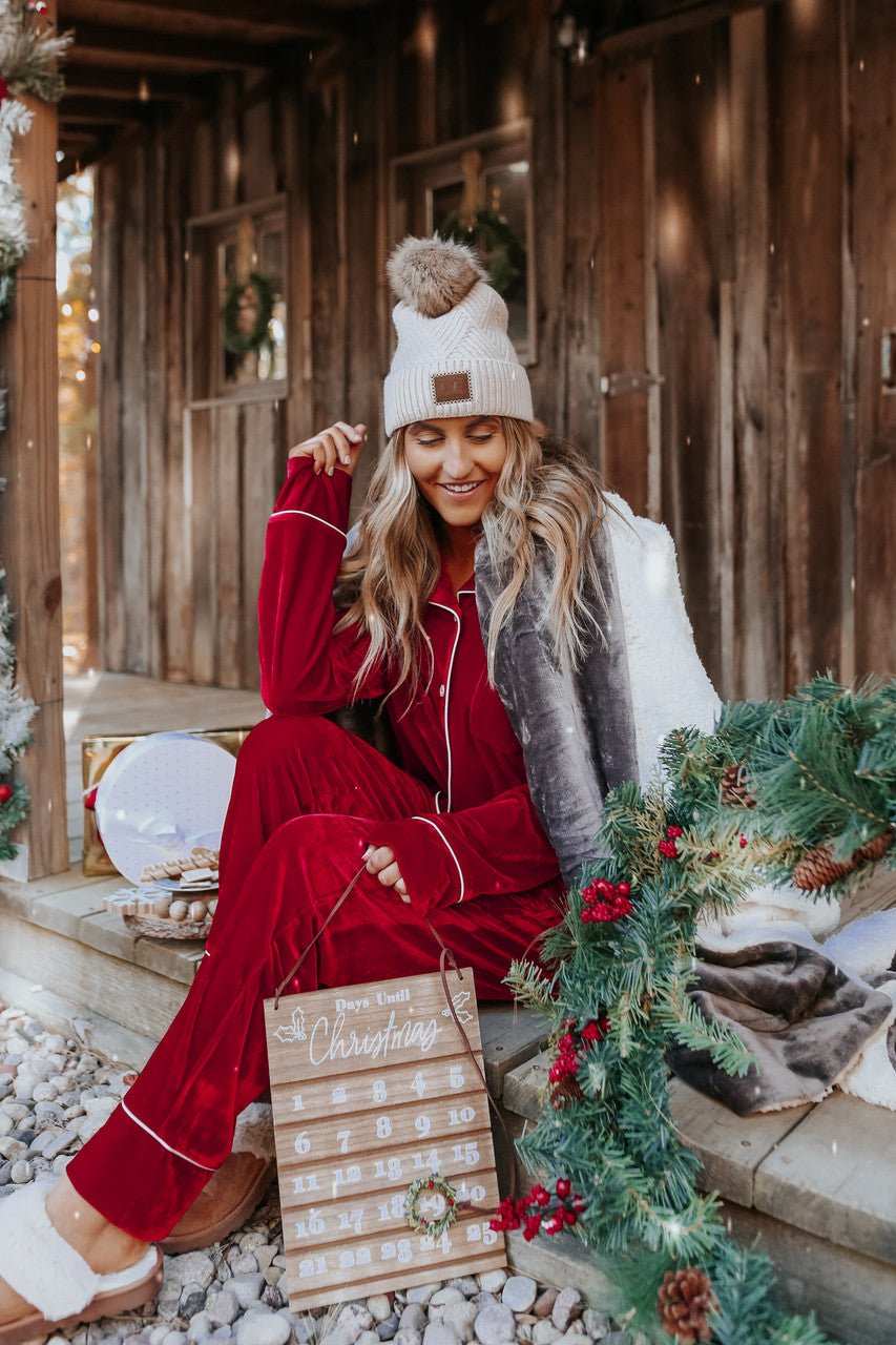Woman in a North Pole Burgundy Velvet Pajama Set sits on a festive porch holding a Christmas countdown calendar.