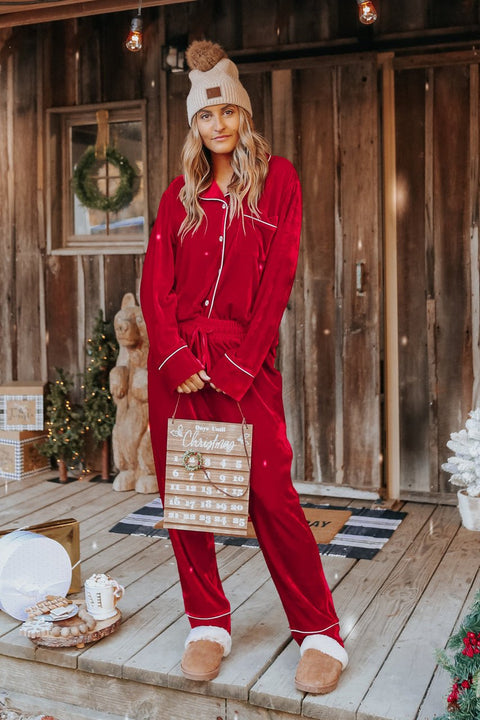 Woman in North Pole Burgundy Velvet Pajama Set stands on a rustic porch, holding a Christmas sign amid festive holiday decor.