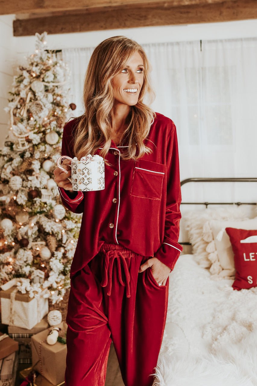 A woman in the North Pole Burgundy Velvet Pajama Set smiles with a mug by a Christmas tree and cozy bed.