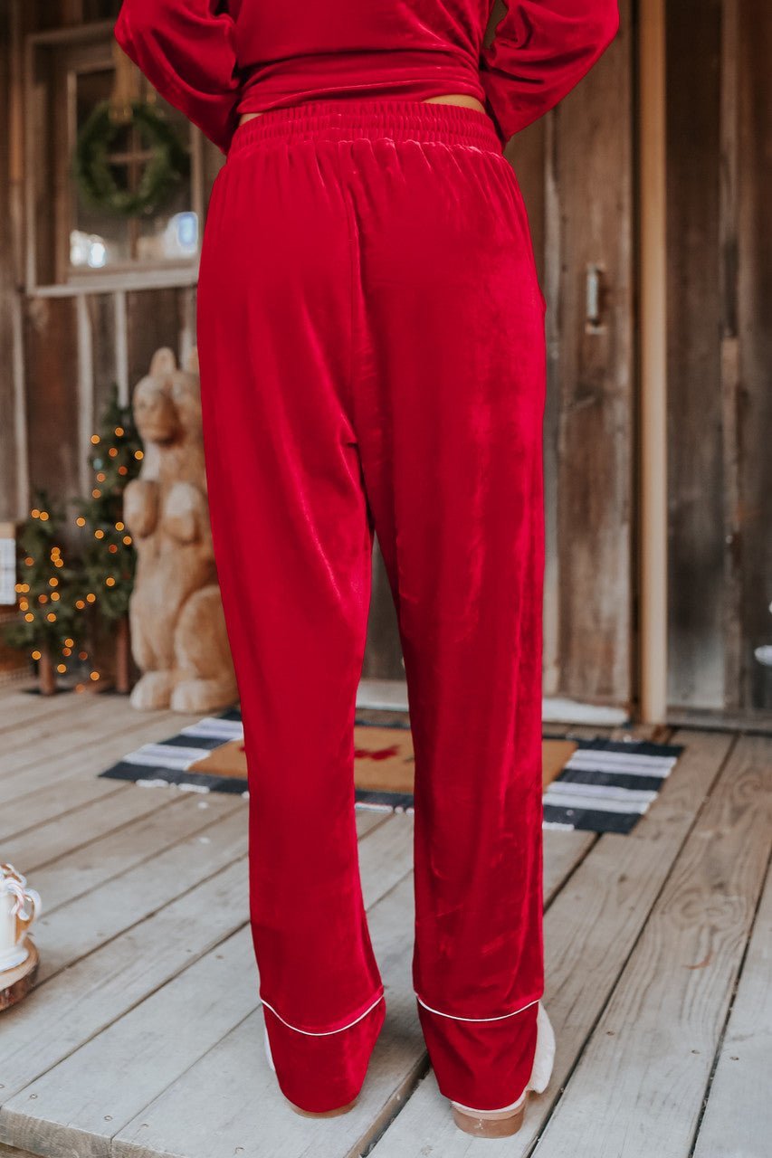 Person wears the North Pole Burgundy Velvet Pajama Set - FINAL SALE, standing on a holiday-decorated wooden porch.