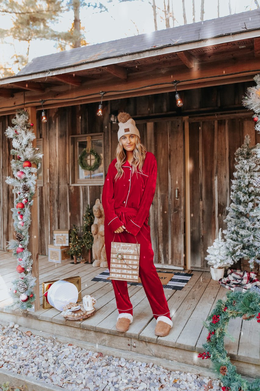 A woman in the North Pole Burgundy Velvet Pajama Set stands on a festive porch, holding a holiday sign among Christmas decorations.