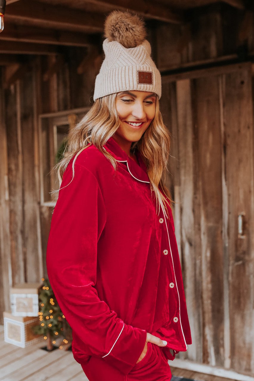 Woman wearing the North Pole Burgundy Velvet Pajama Set and a knit pom-pom hat, smiling outside by a rustic wooden building.