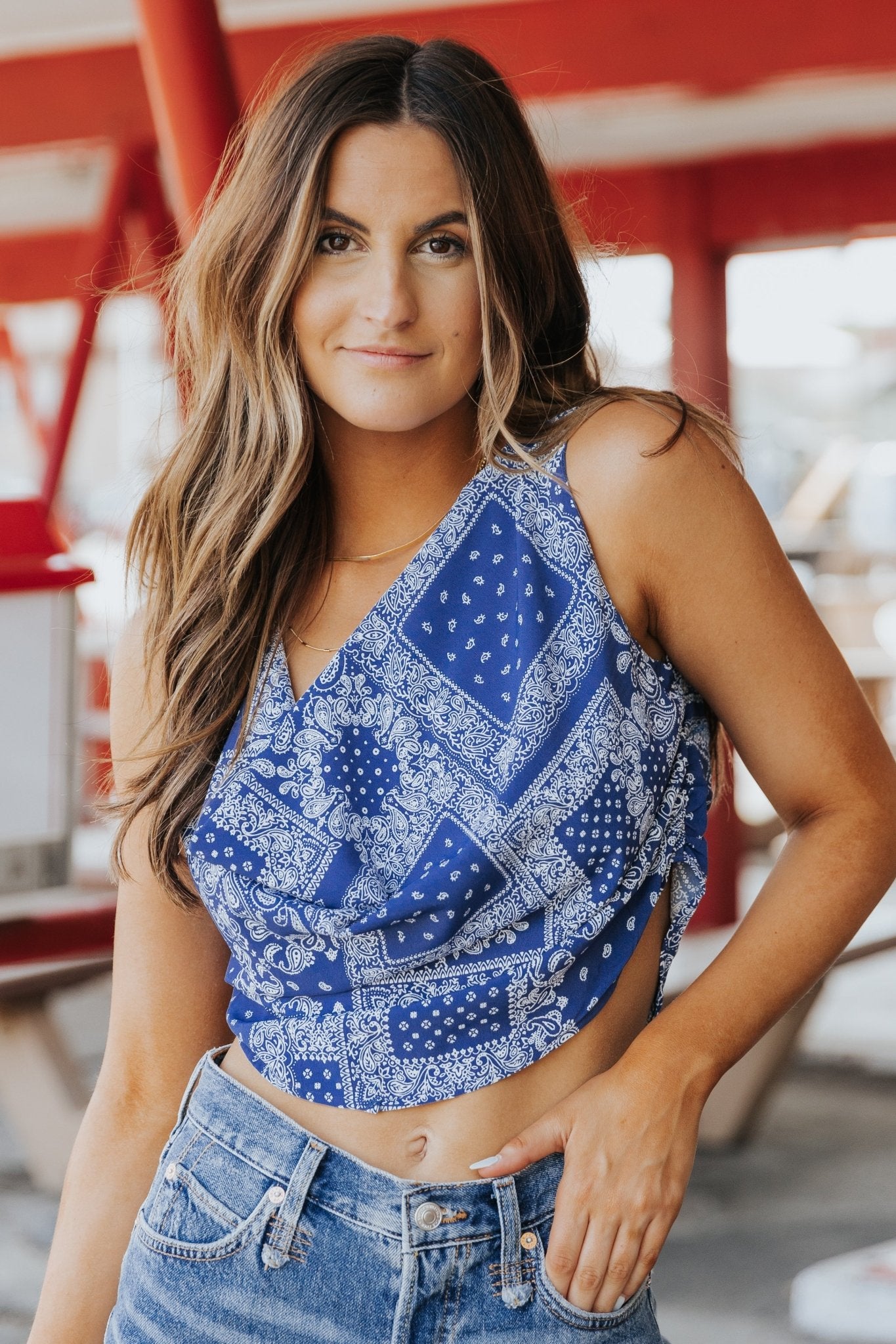 Woman with long brown hair wears the Out West Blue Bandana Print Sleeveless Cowl Neck Top, smiling outdoors.